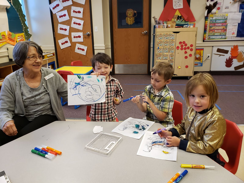 an adult with three young children drawing with markers; one of them is grinning and holding up their artwork