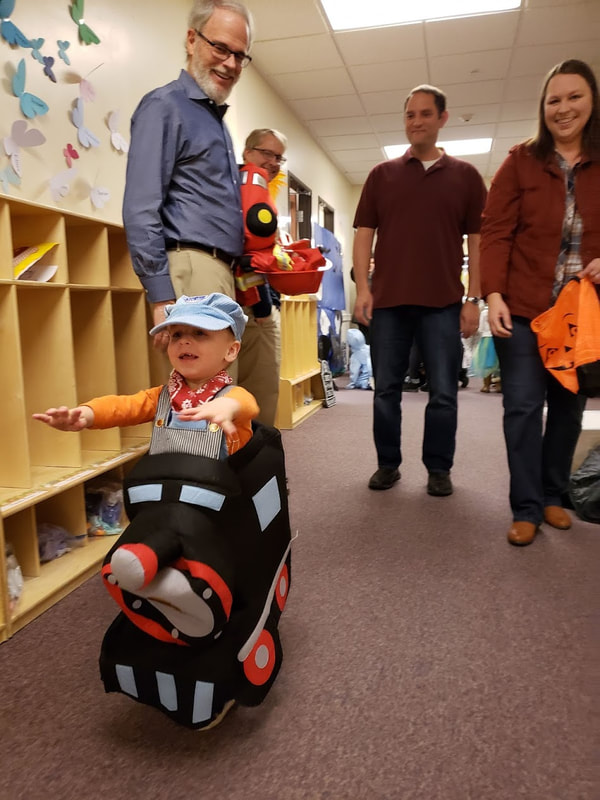 a group of adults grinning at a toddler dressed as a train-and-conductor, in a hallway of trick-or-treaters